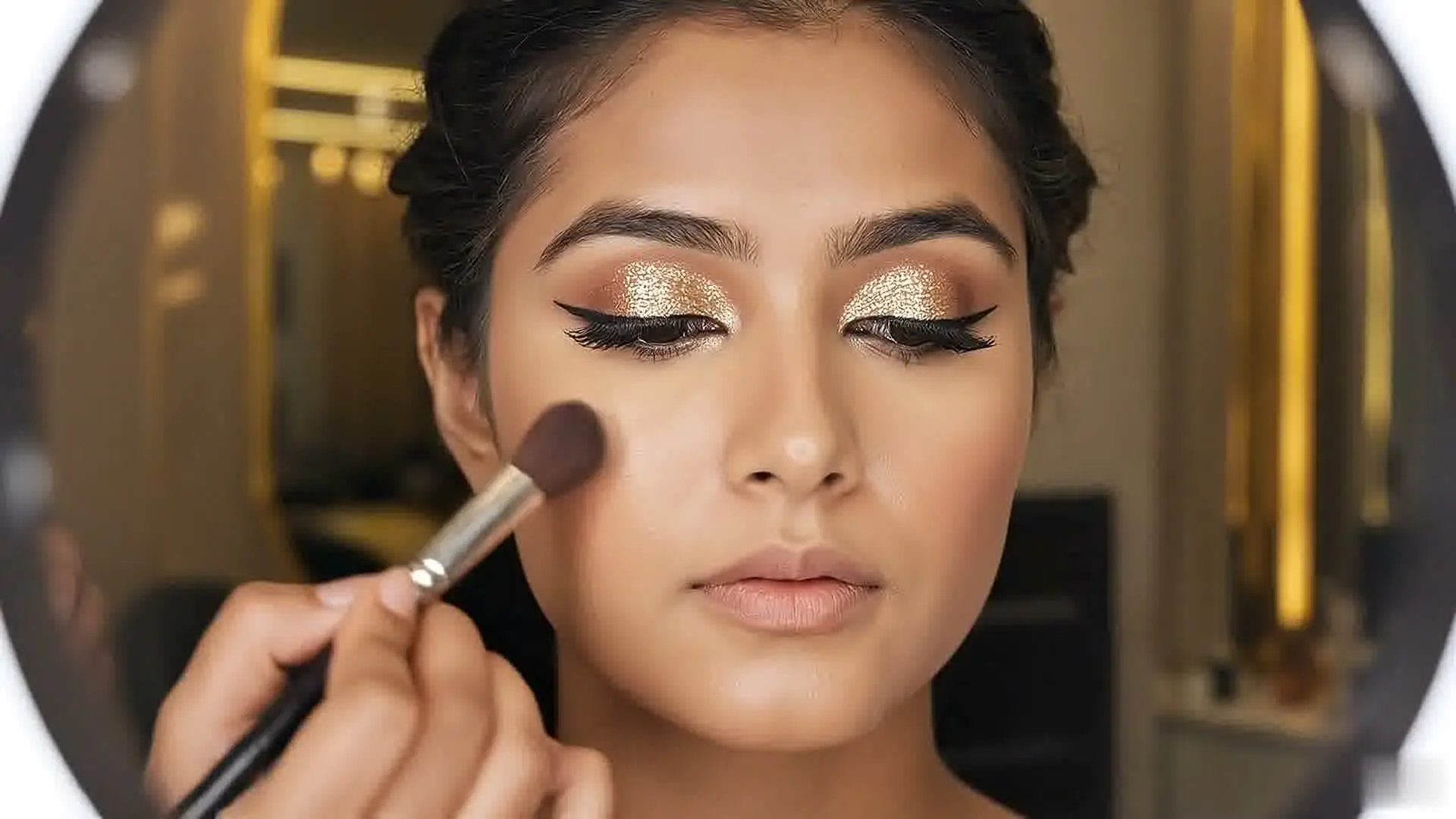 A woman with gold glitter eyeshadow and winged liner as a brush sweeps across her cheek